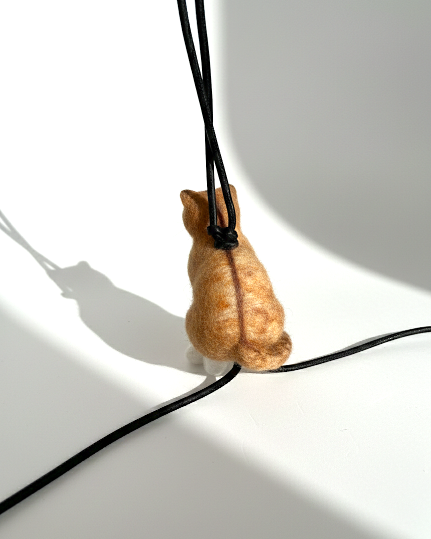 Back view of Merino wool needle-felted cat-shaped pendant on a black lambskin cord against a white background.