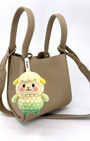 A taupe, textured leather mini tote bag with two top handles and a shoulder strap is shown against a plain background. Hanging from the front handle is a small, needle-felted sheep keychain. The sheep has a creamy white bubbly wool body and head, a light green lower body, small yellow ears, and pink cheeks. It is attached with a white tag that reads "Woobbi".