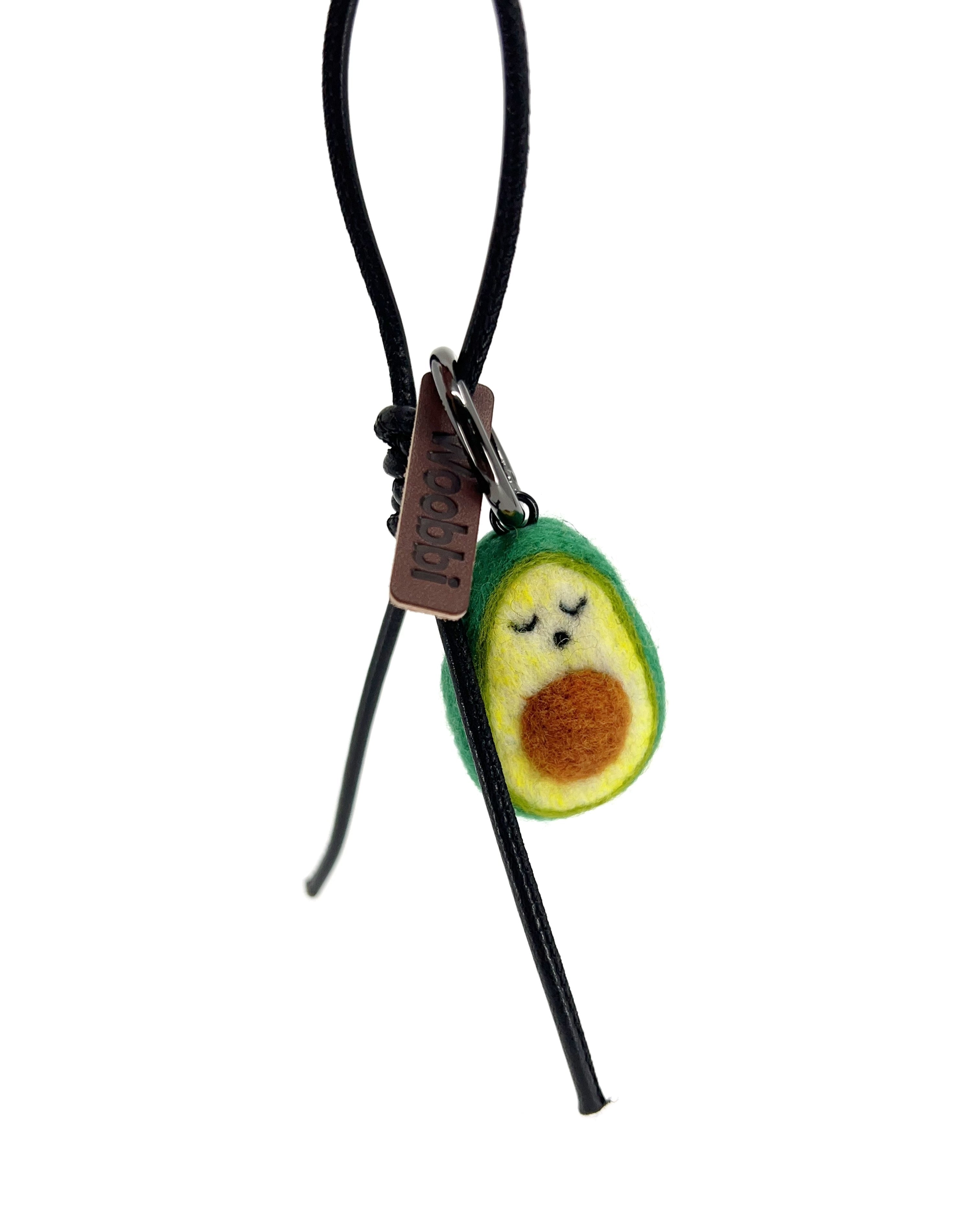 Studio product shot of needle-felted avocado bag charm with red leather cord and brown Woobbi brand tag, isolated on a white background.