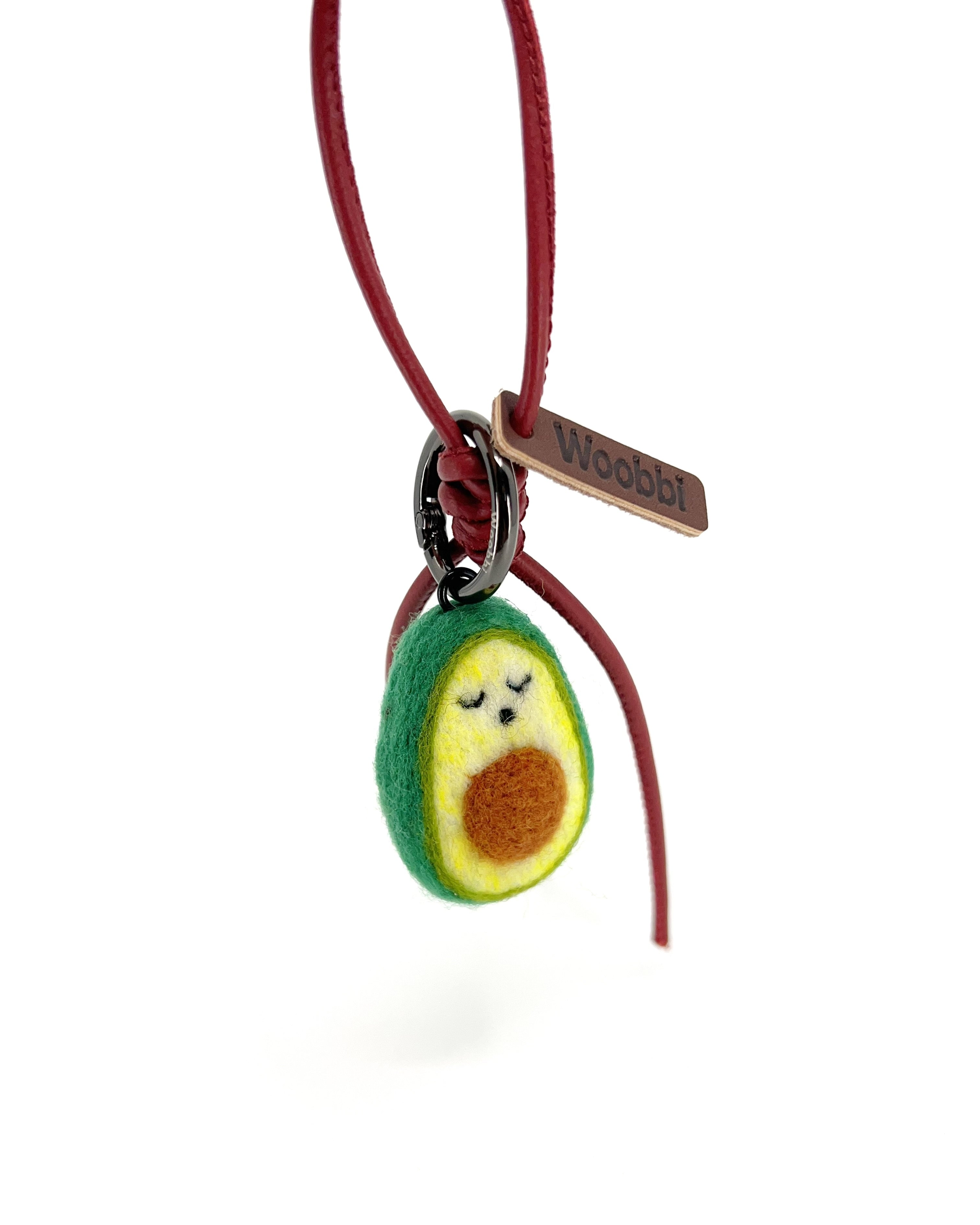 Studio product shot of needle-felted avocado bag charm with red leather cord and brown Woobbi brand tag, isolated on a white background.