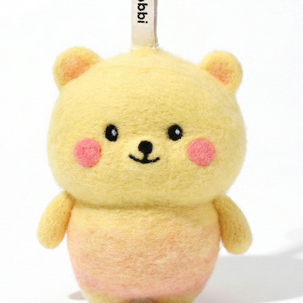 Front view of a handmade Woobbi bear keychain, crafted from yellow and pink bubbly wool felt. It has small yellow ears, rosy pink cheeks, and a smiling face with black eyes and nose. A small tag with the brand name 'Woobbi' is attached to the top. It is set against a plain white background.