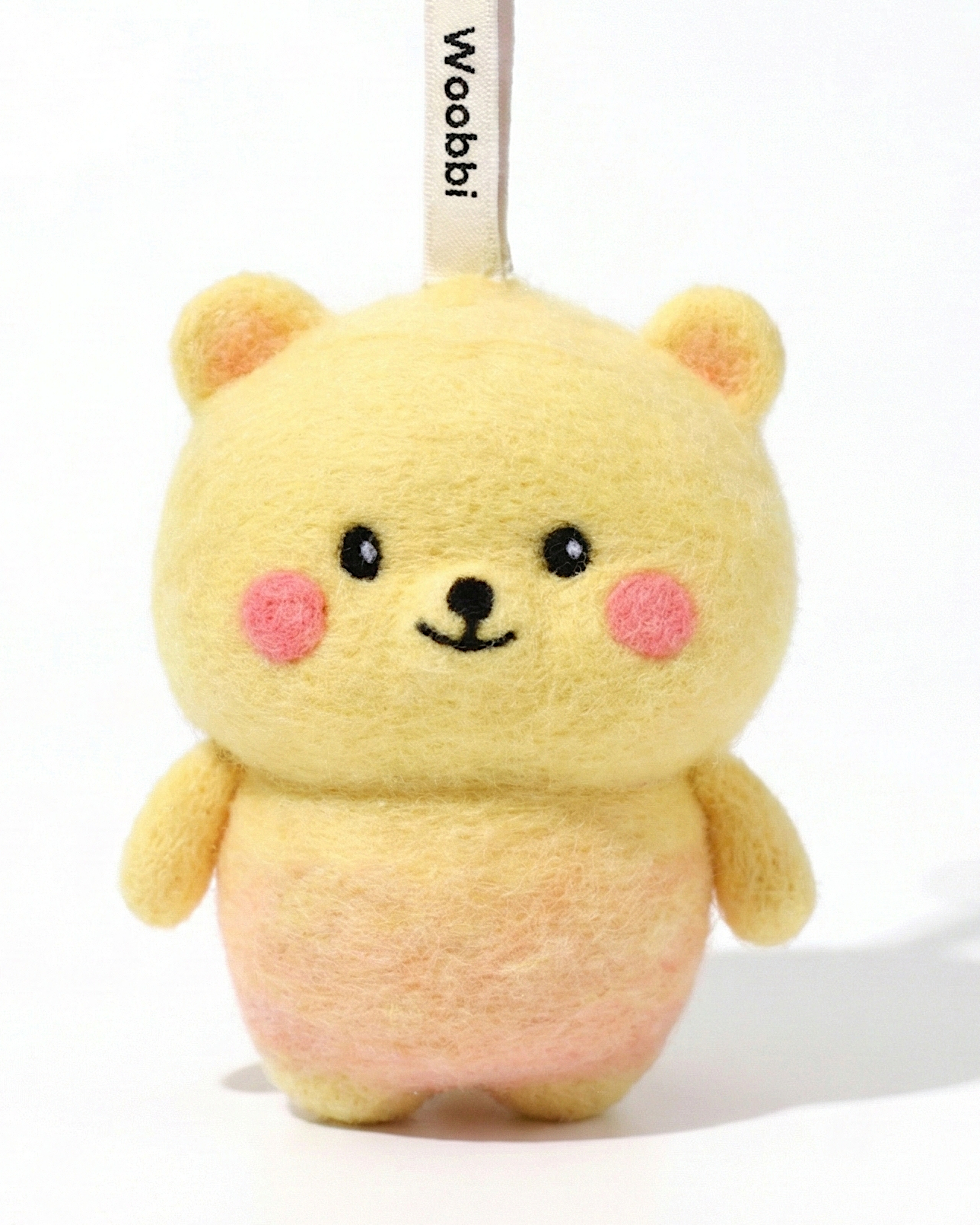 Front view of a handmade Woobbi bear keychain, crafted from yellow and pink bubbly wool felt. It has small yellow ears, rosy pink cheeks, and a smiling face with black eyes and nose. A small tag with the brand name 'Woobbi' is attached to the top. It is set against a plain white background.