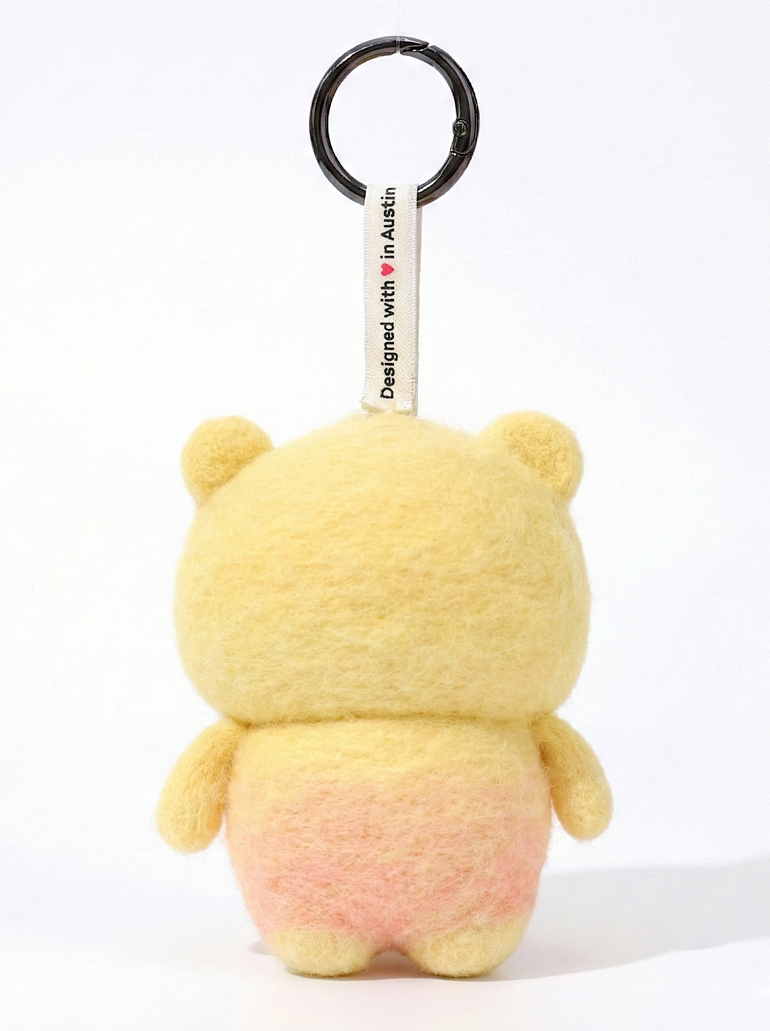 Back view of a handmade Woobbi bear keychain, made from yellow and pink bubbly wool felt. It features a small tag that says 'Designed with ❤️ in Austin' and a round black metal keyring. The sheep has small yellow ears. It is set against a plain white background.