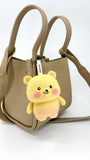 A taupe, textured leather mini tote bag with two top handles and a shoulder strap is shown against a plain background. Hanging from the front handle is a small, needle-felted bear keychain. The bear has a yellow bubbly wool body and head, a light pink lower body, small yellow ears, and pink cheeks. It is attached with a white tag that reads "Woobbi".