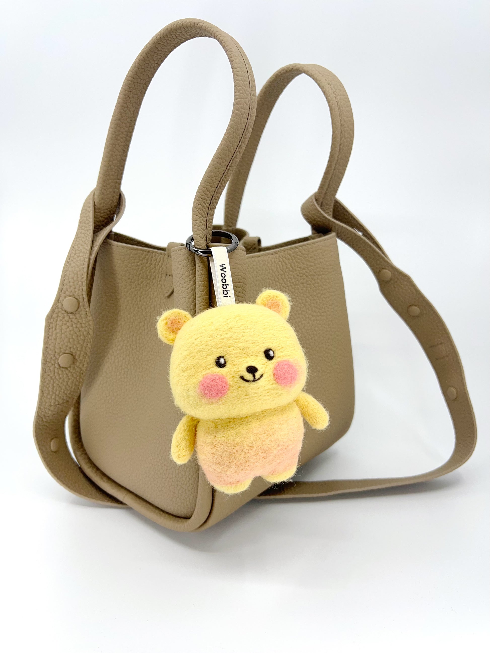 A taupe, textured leather mini tote bag with two top handles and a shoulder strap is shown against a plain background. Hanging from the front handle is a small, needle-felted bear keychain. The bear has a yellow bubbly wool body and head, a light pink lower body, small yellow ears, and pink cheeks. It is attached with a white tag that reads "Woobbi".