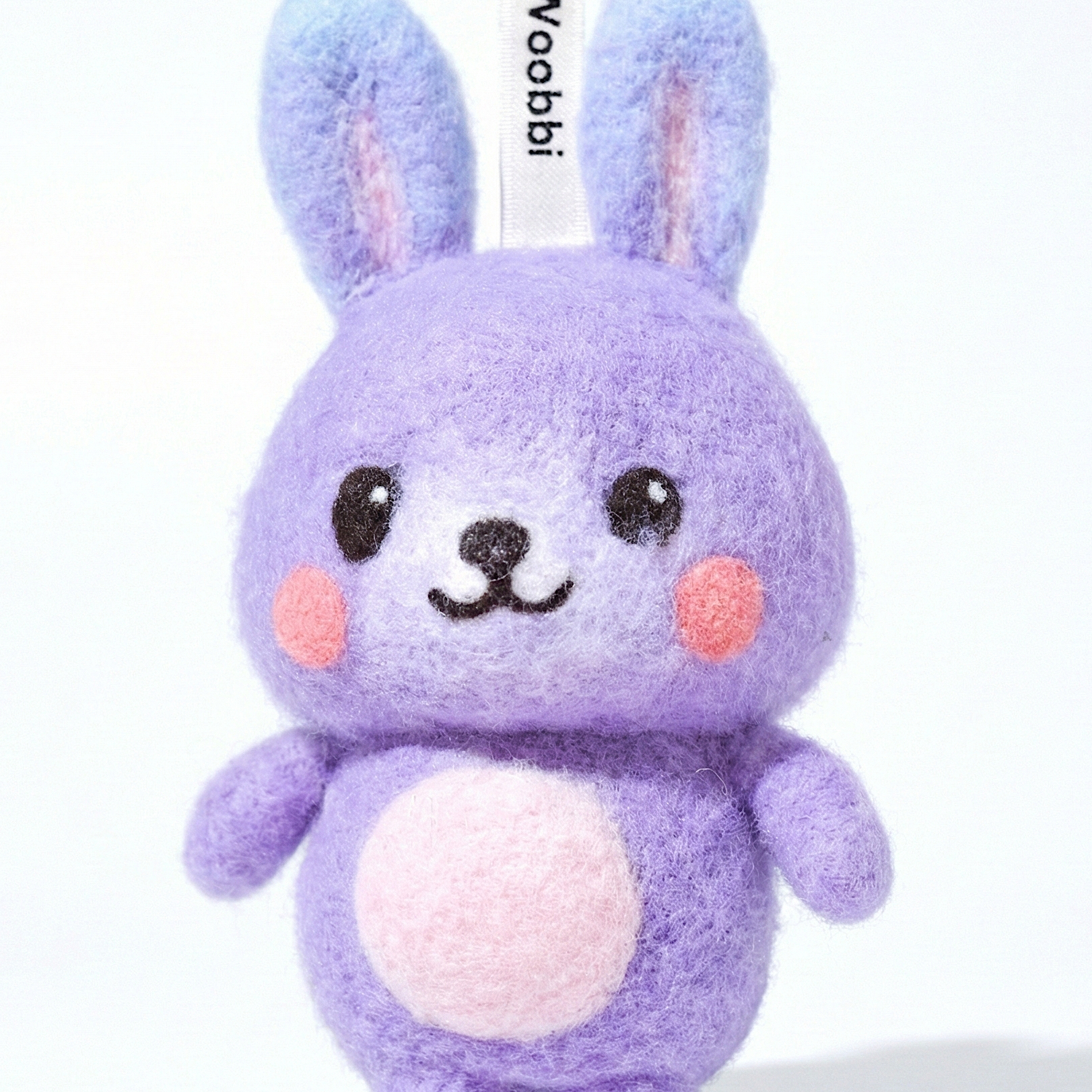 Front view of a handmade Woobbi bunny keychain, crafted from purple bubbly wool felt. It has small yellow ears, rosy pink cheeks, and a smiling face with black eyes and nose. A small tag with the brand name 'Woobbi' is attached to the top. It is set against a plain white background.