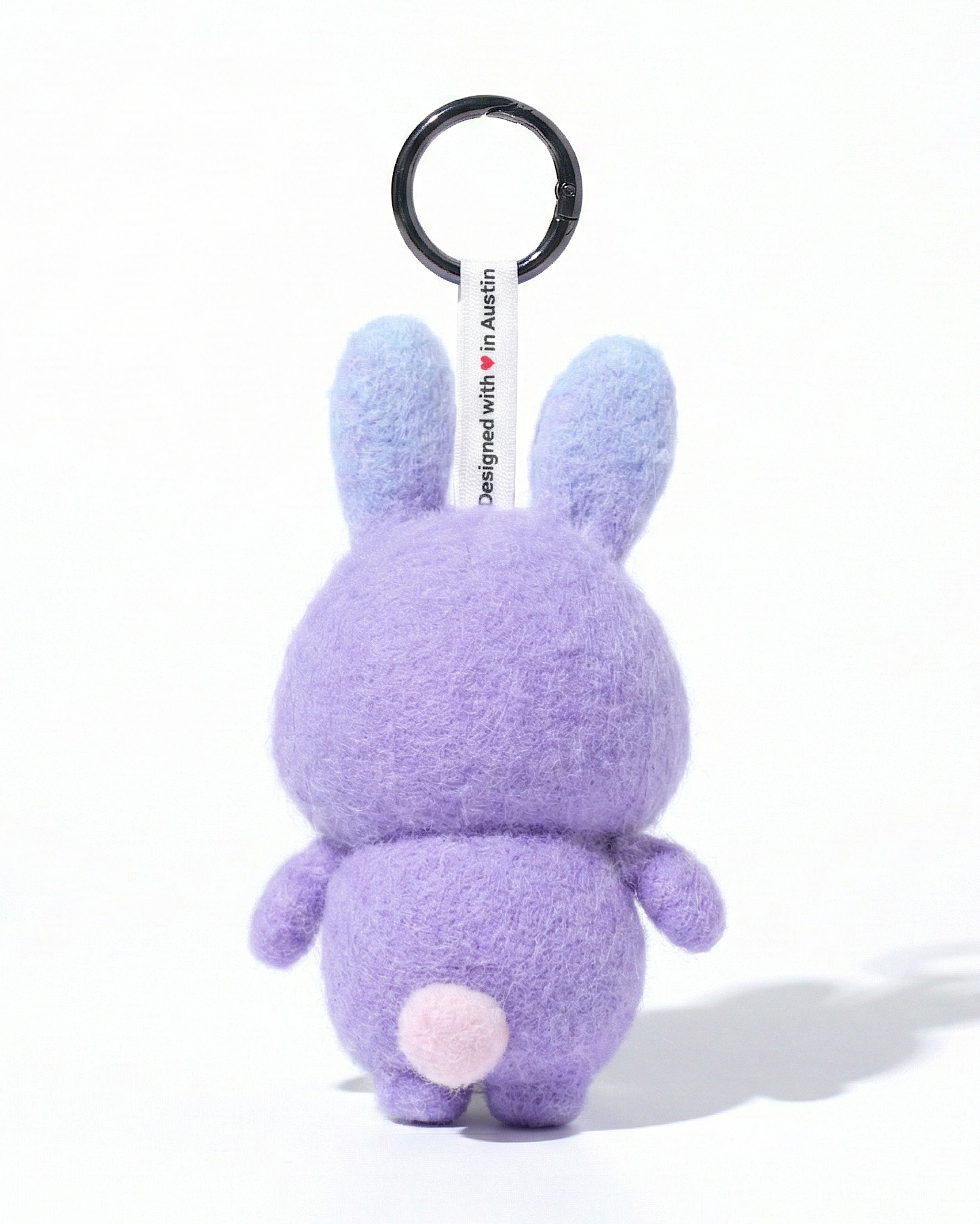 Back view of a handmade Woobbi bunny keychain, made from purple bubbly wool felt. It features a small tag that says 'Designed with ❤️ in Austin' and a round black metal keyring. The sheep has long blue ears and a tiny white tail. It is set against a plain white background.