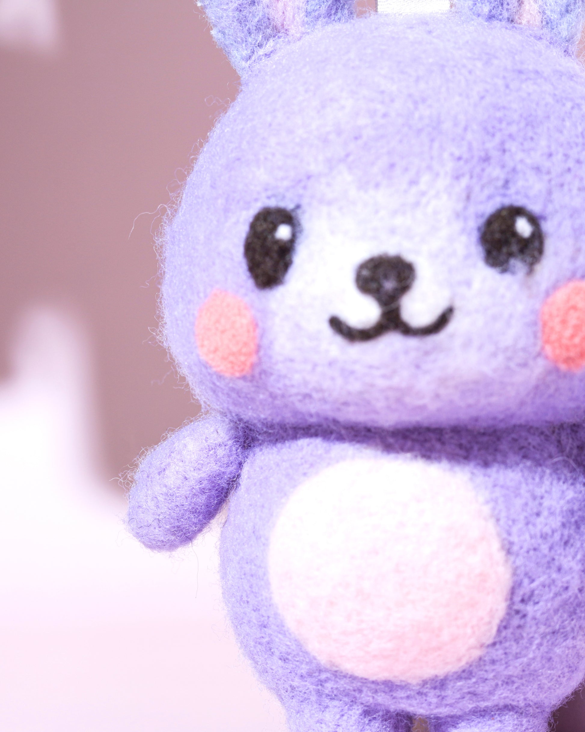 Macro close-up of purple needle-felted bunny face showing soft Merino wool texture, pink cheeks, and hand-stitched smile.