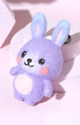 High-angle view of cute purple wool rabbit bag charm with blue-tipped ears and pink belly, displayed on a modern pink pedestal.
