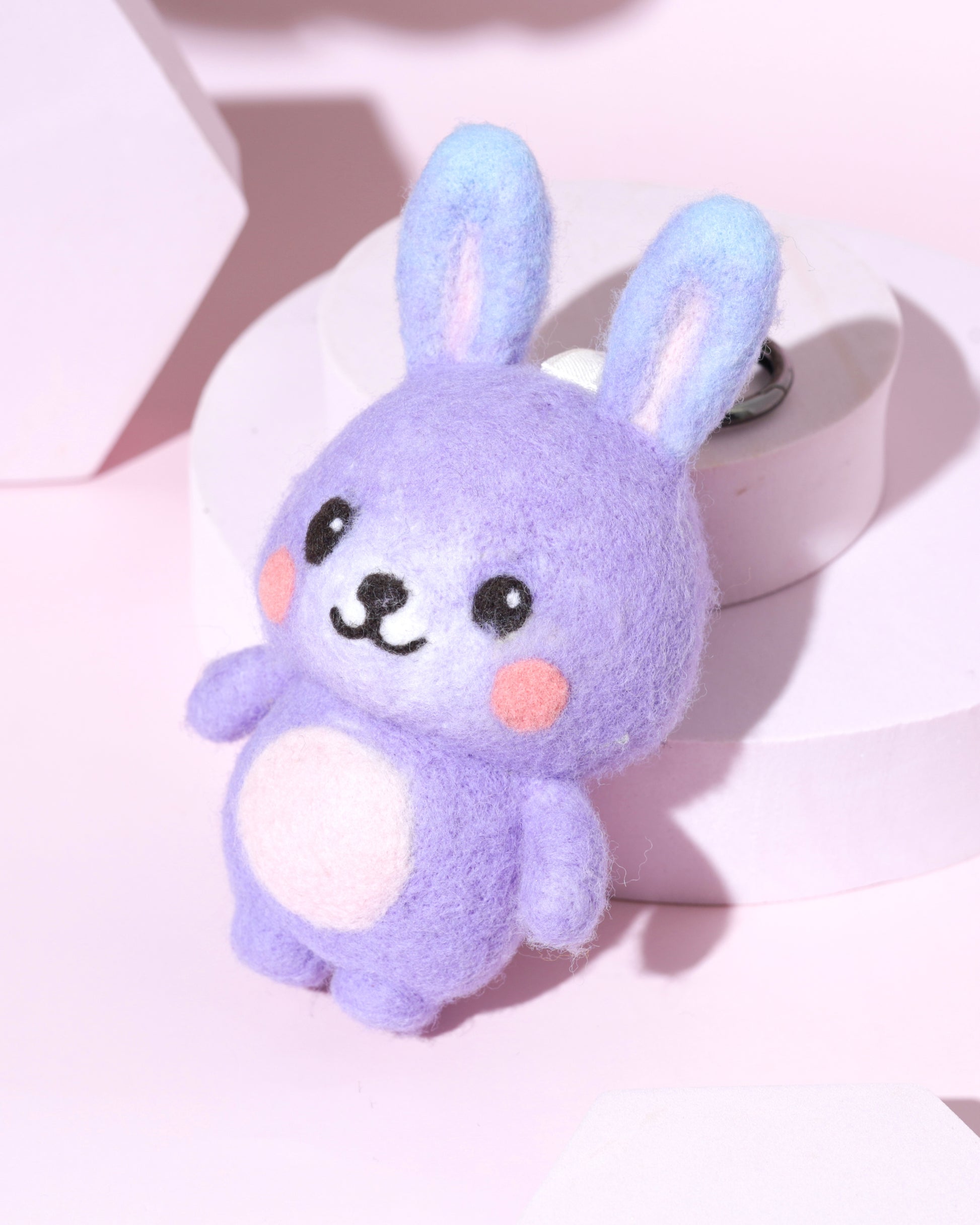 High-angle view of cute purple wool rabbit bag charm with blue-tipped ears and pink belly, displayed on a modern pink pedestal.
