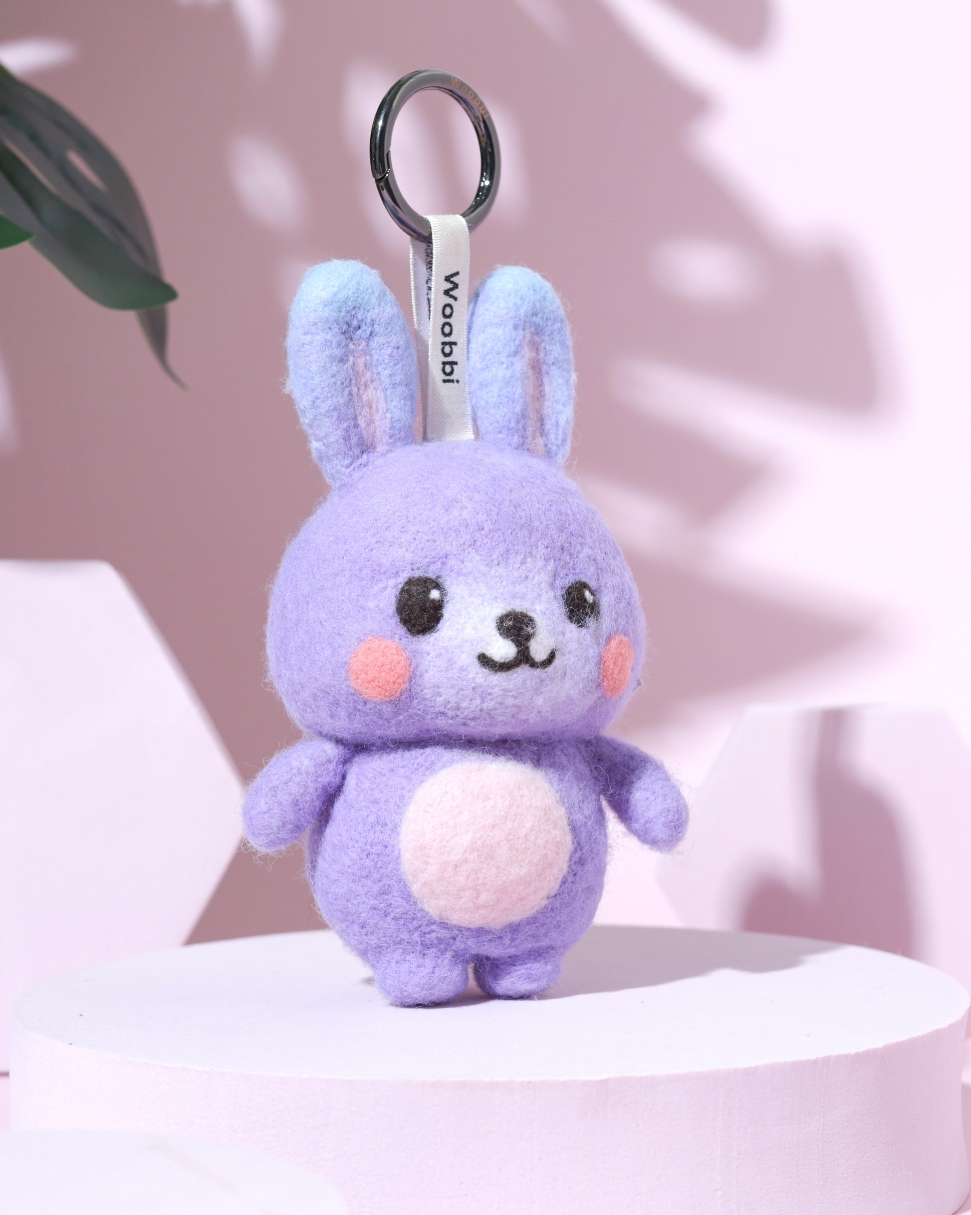 Front view of handcrafted needle-felted purple bunny keychain standing on a pink platform, featuring a durable black metal ring and white Woobbi brand tag.