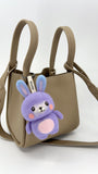 A taupe, textured leather mini tote bag with two top handles and a shoulder strap is shown against a plain background. Hanging from the front handle is a small, needle-felted bunny keychain. The bunny has a purple bubbly wool body and head, long purple ears, and pink cheeks. It is attached with a white tag that reads "Woobbi".