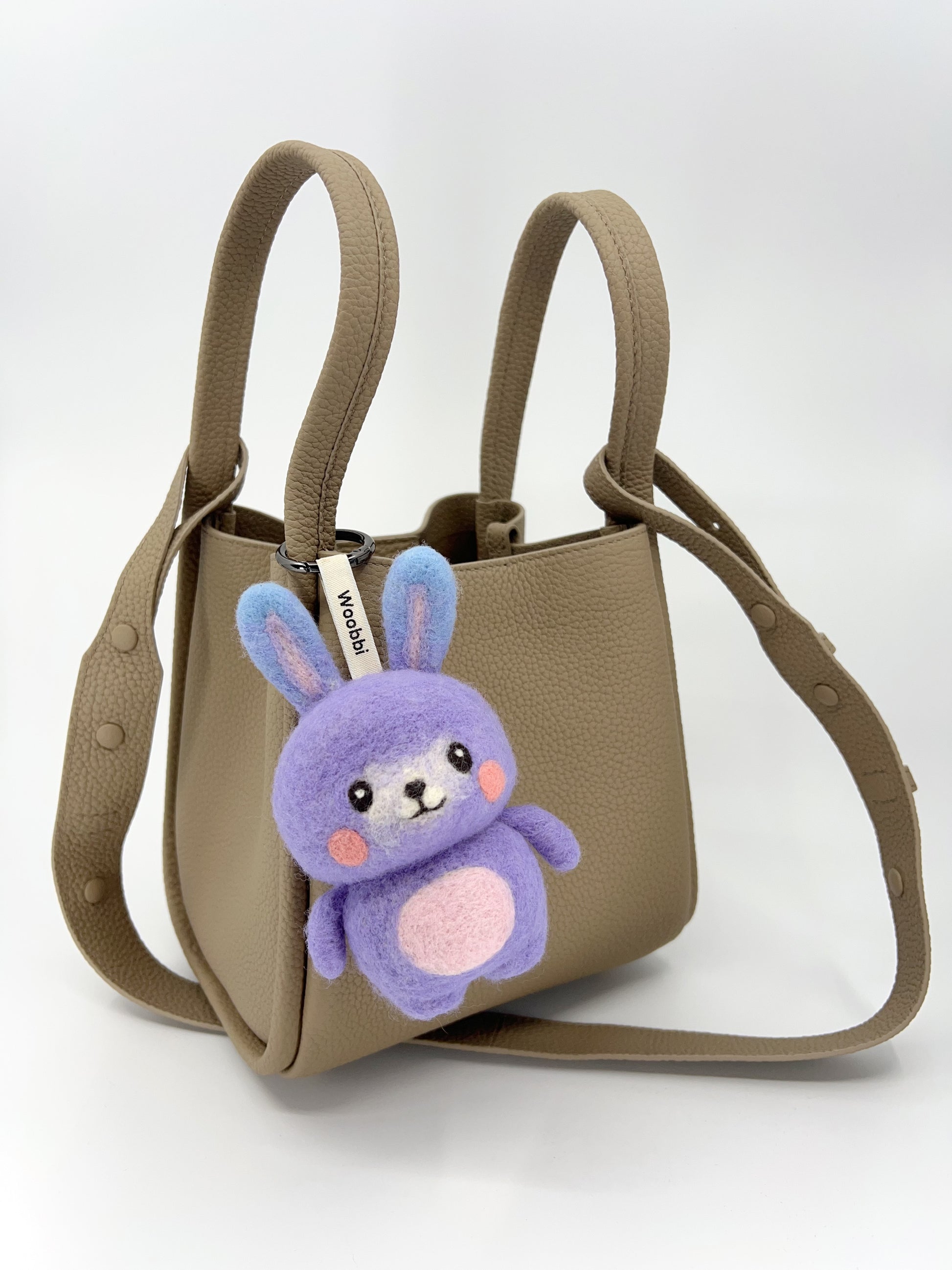 A taupe, textured leather mini tote bag with two top handles and a shoulder strap is shown against a plain background. Hanging from the front handle is a small, needle-felted bunny keychain. The bunny has a purple bubbly wool body and head, long purple ears, and pink cheeks. It is attached with a white tag that reads "Woobbi".