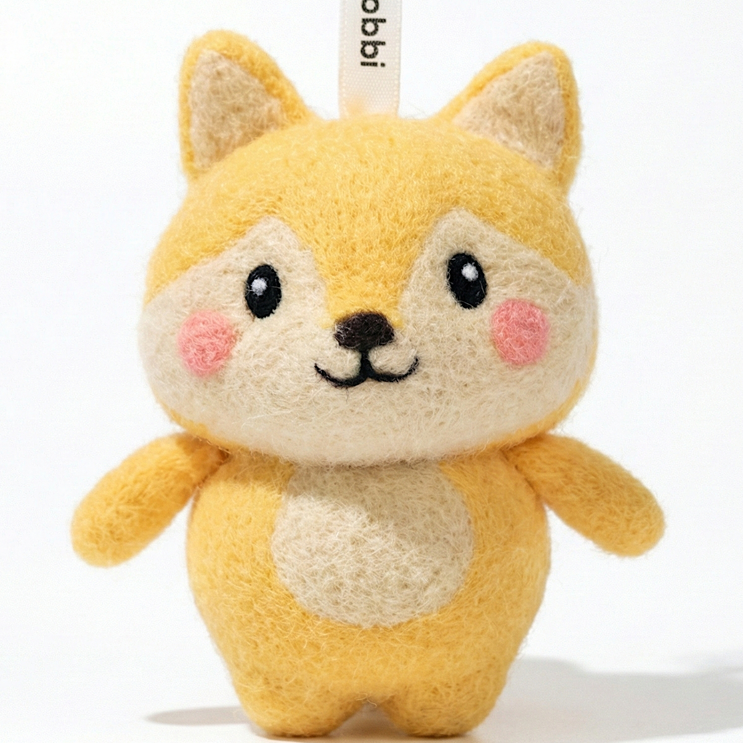 Front view of a handmade Woobbi fox keychain, made from soft yellow and cream wool felt. The fox has pointed ears, rosy pink cheeks, and a happy smiling face with black eyes and nose. It features a white belly patch and is attached to a black metal keyring with a tag labeled 'Woobbi'. The background is plain white.