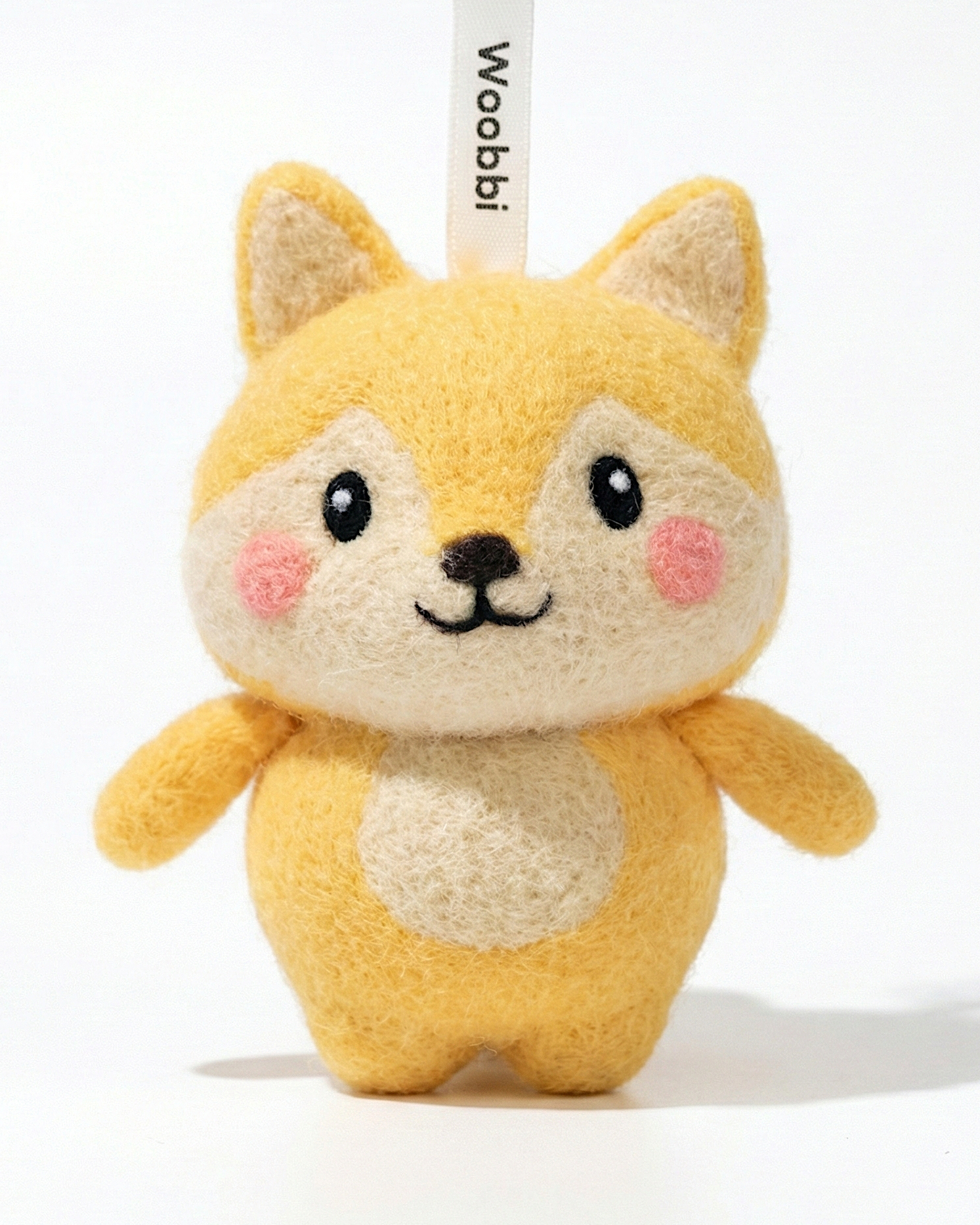Front view of a handmade Woobbi fox keychain, made from soft yellow and cream wool felt. The fox has pointed ears, rosy pink cheeks, and a happy smiling face with black eyes and nose. It features a white belly patch and is attached to a black metal keyring with a tag labeled 'Woobbi'. The background is plain white.