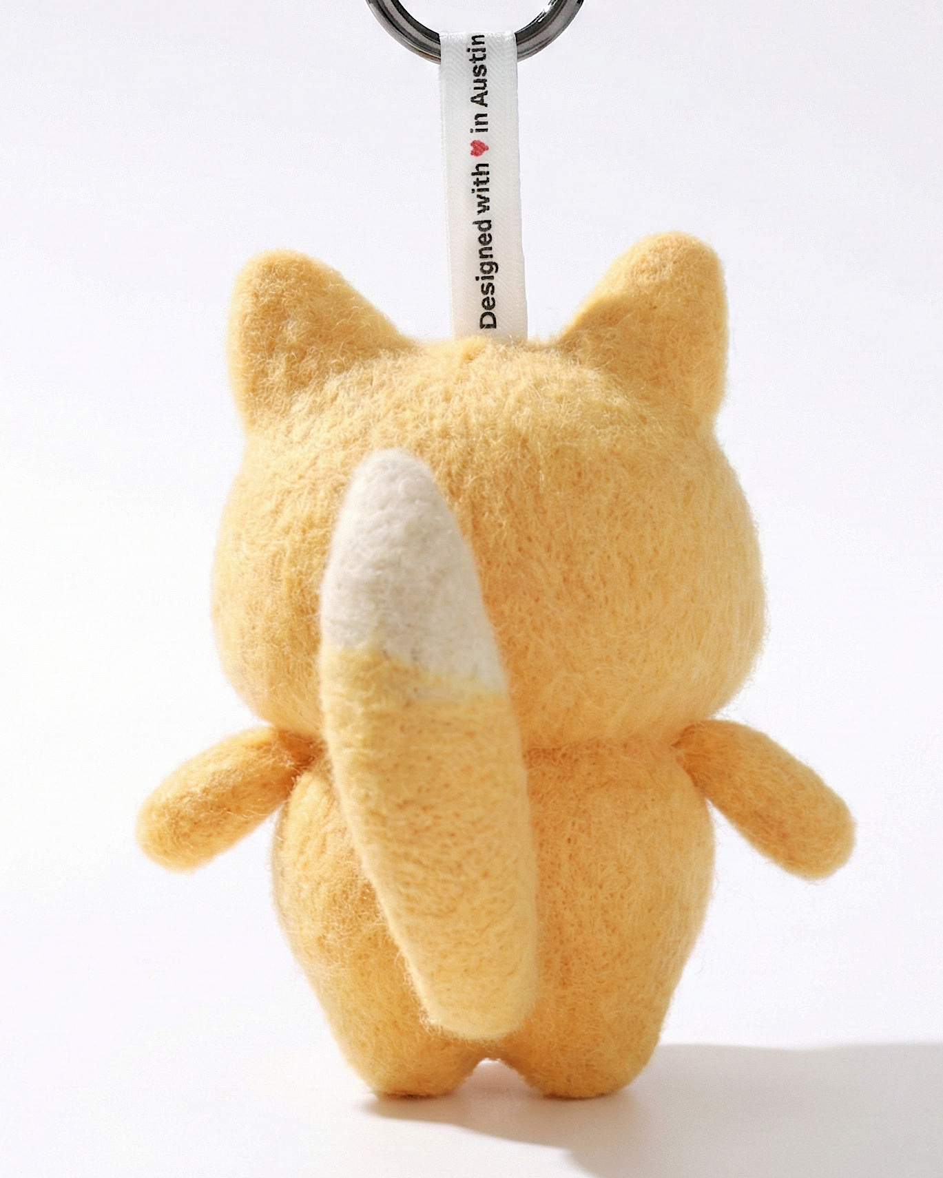 Back view of a handmade Woobbi fox keychain, crafted from fuzzy yellow wool felt. It features a fluffy tail with a white tip and a small white tag attached to the top that reads 'Designed with ❤️ in Austin'. The keychain has a round black metal ring and is set against a plain white background.