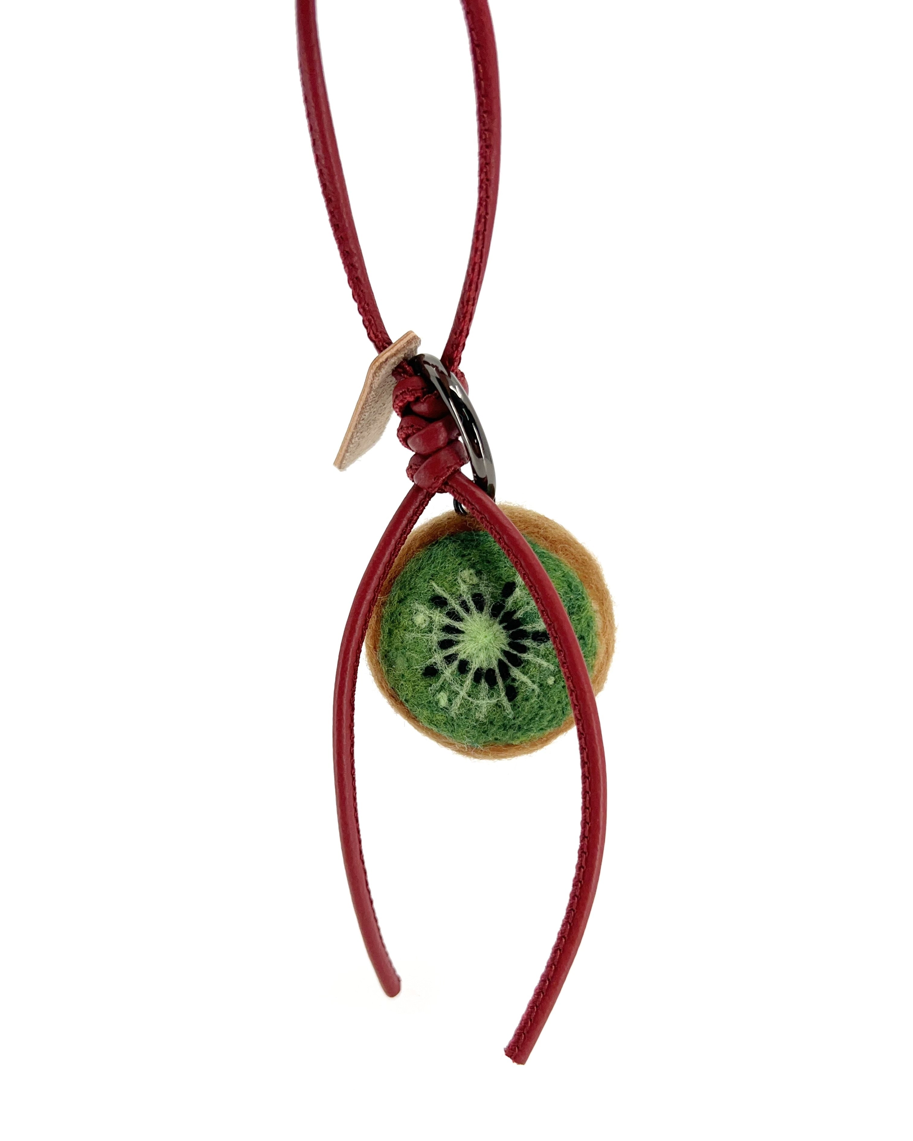 Back view of needle-felted kiwi slice showing bright green fruit detail and black seeds, attached to a deep red leather strap.