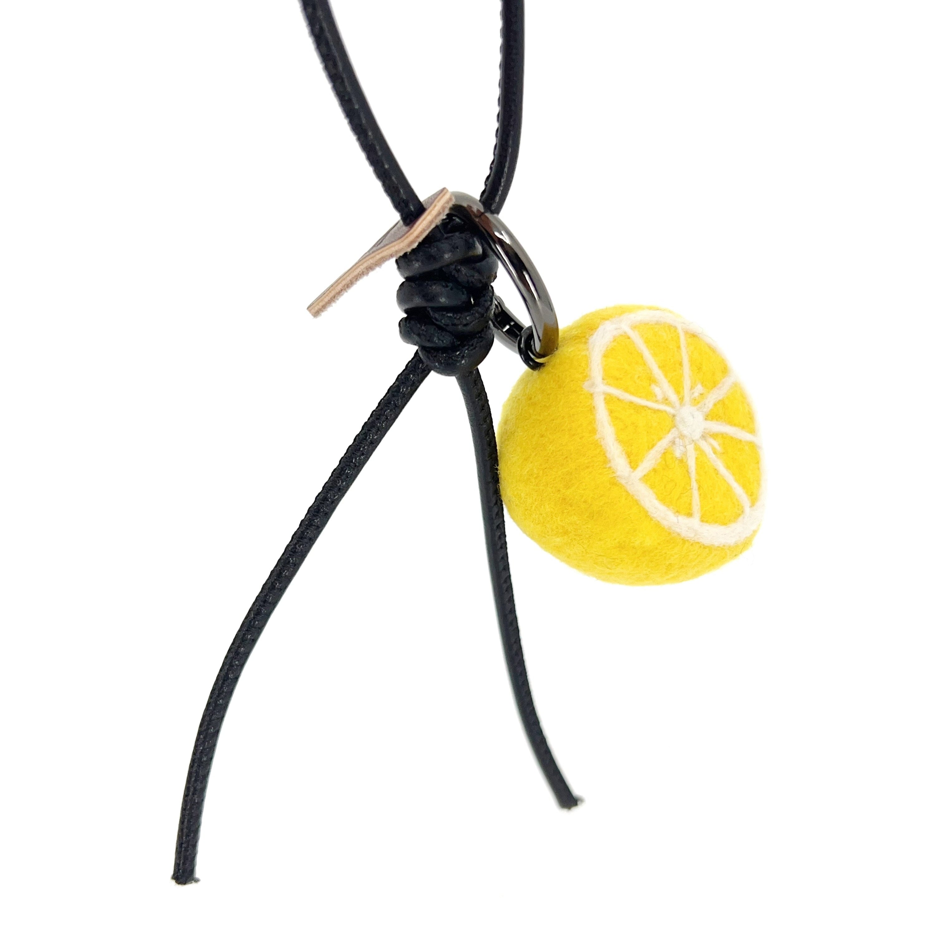 Hanging view of needle-felted lemon slice bag charm with black leather cord, showing detailed white pith fiber art and brown brand tag.