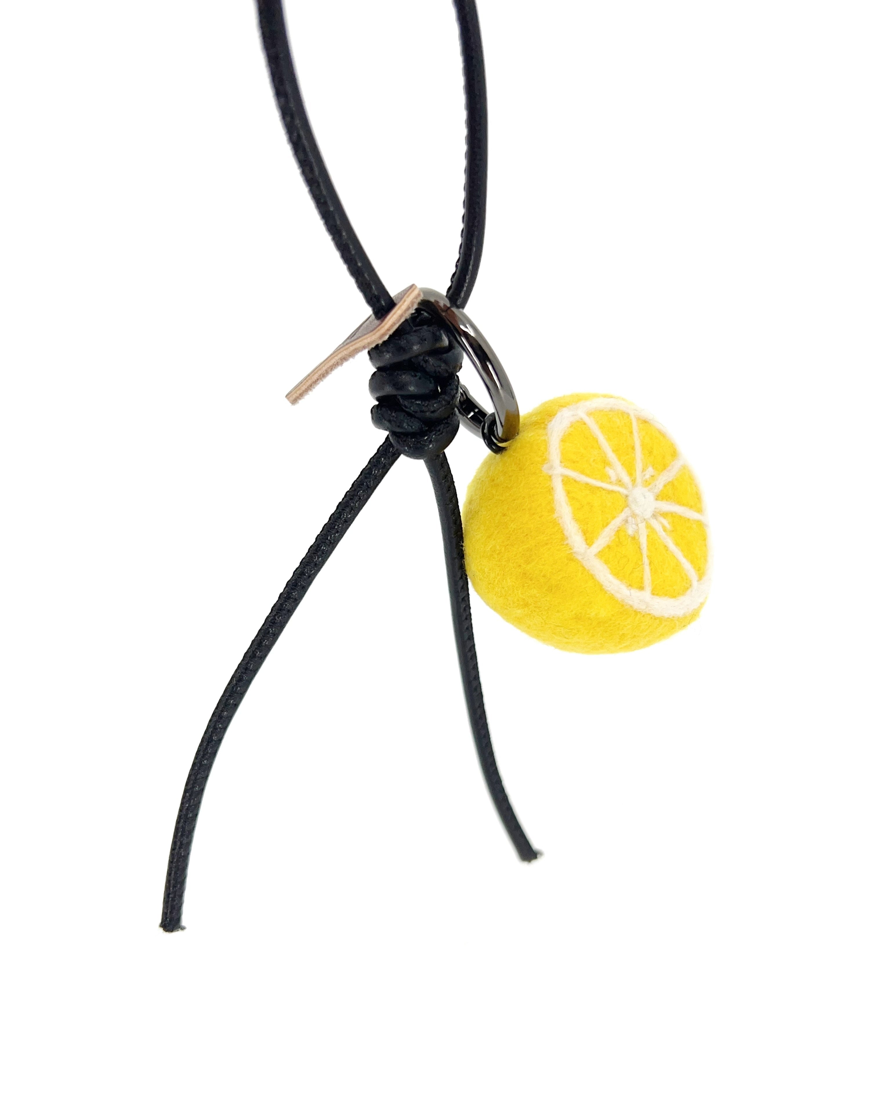 Hanging view of needle-felted lemon slice bag charm with black leather cord, showing detailed white pith fiber art and brown brand tag.