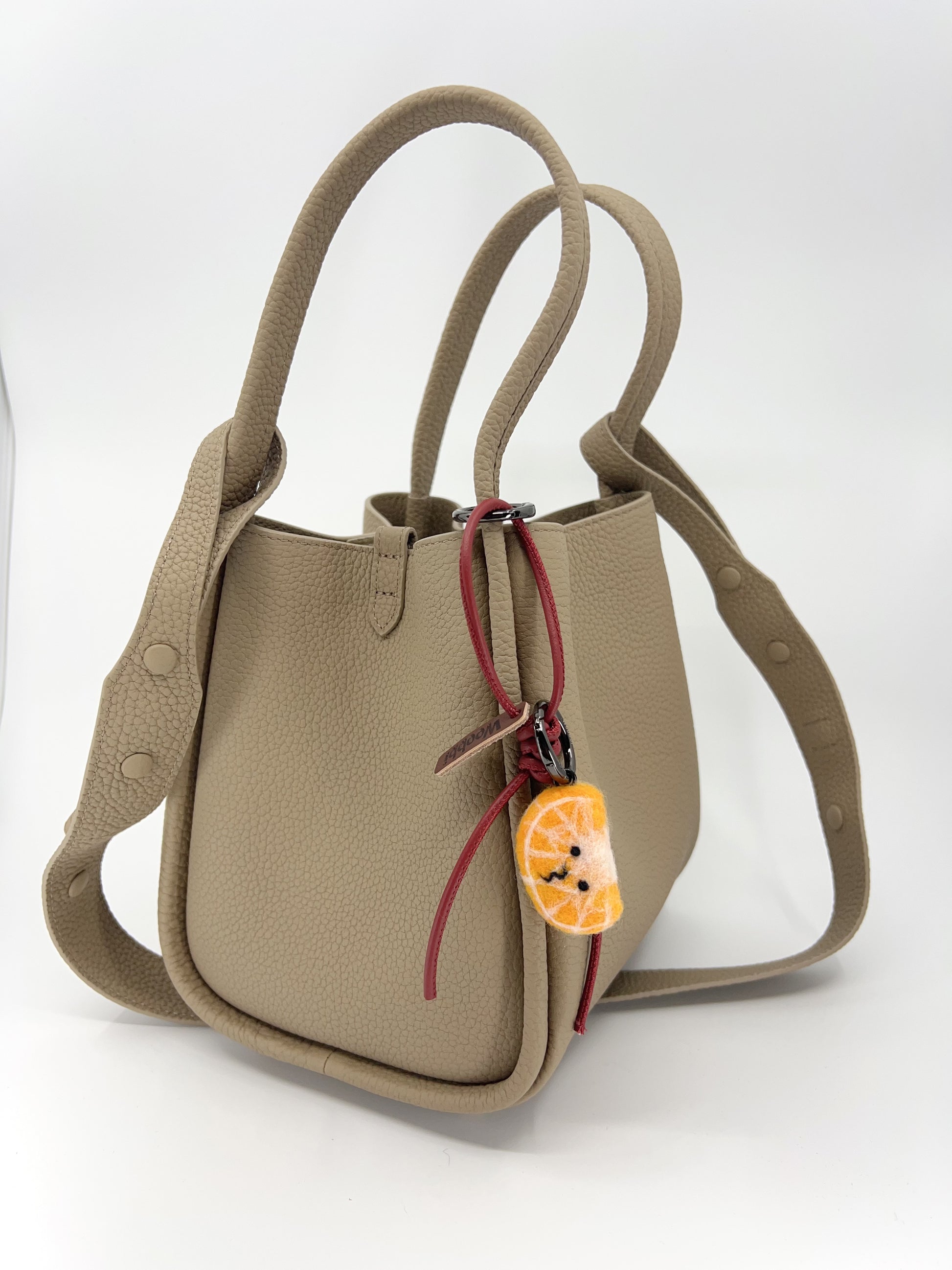 Lifestyle shot of handcrafted needle-felted orange slice bag charm with red leather strap clipped onto a beige leather handbag, showing the cute fruit accessory in use.