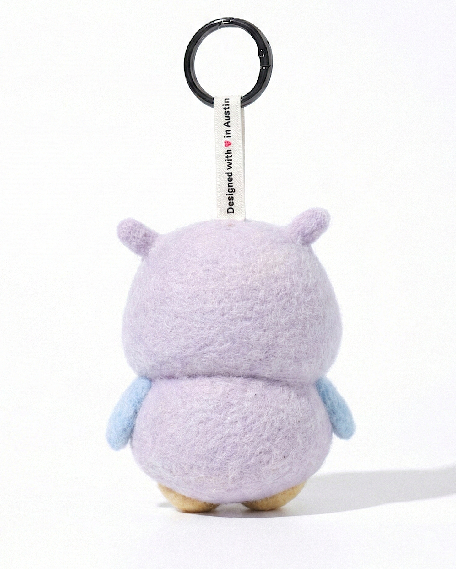 Back view of a handmade Woobbi owl keychain, showing its rounded lavender wool felt body and small pointed ears. The keychain features light blue wings on the sides and a white tag attached to the top that reads 'Designed with ❤️ in Austin'. A round black metal keyring is connected to the tag. The product is set against a pure white background.