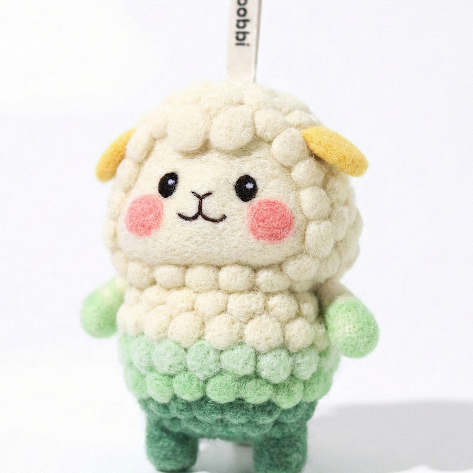Front view of a handmade Woobbi sheep keychain, crafted from off-white and green bubbly wool felt. It has small yellow ears, rosy pink cheeks, and a smiling face with black eyes and nose. A small tag with the brand name 'Woobbi' is attached to the top. It is set against a plain white background.