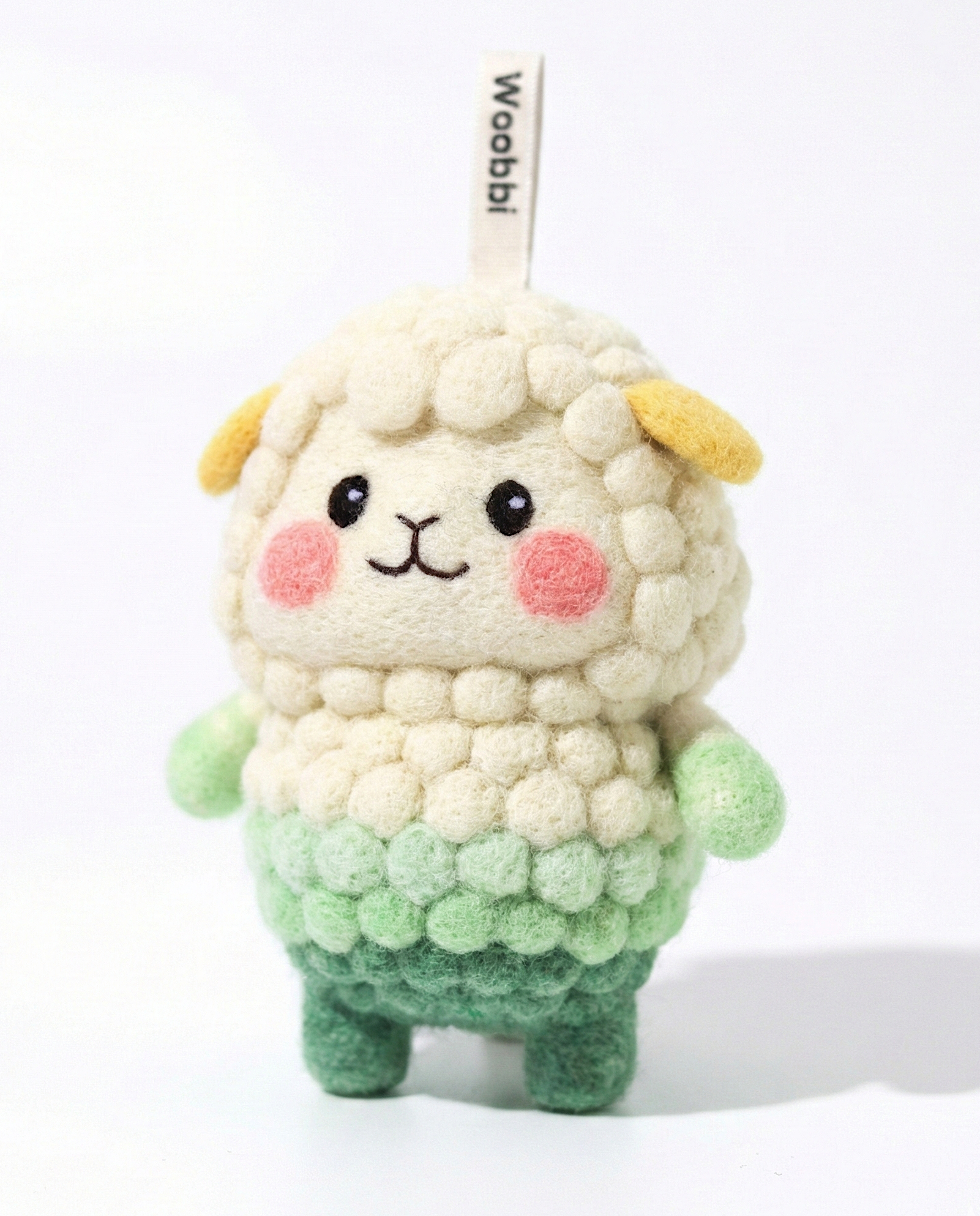 Front view of a handmade Woobbi sheep keychain, crafted from off-white and green bubbly wool felt. It has small yellow ears, rosy pink cheeks, and a smiling face with black eyes and nose. A small tag with the brand name 'Woobbi' is attached to the top. It is set against a plain white background.