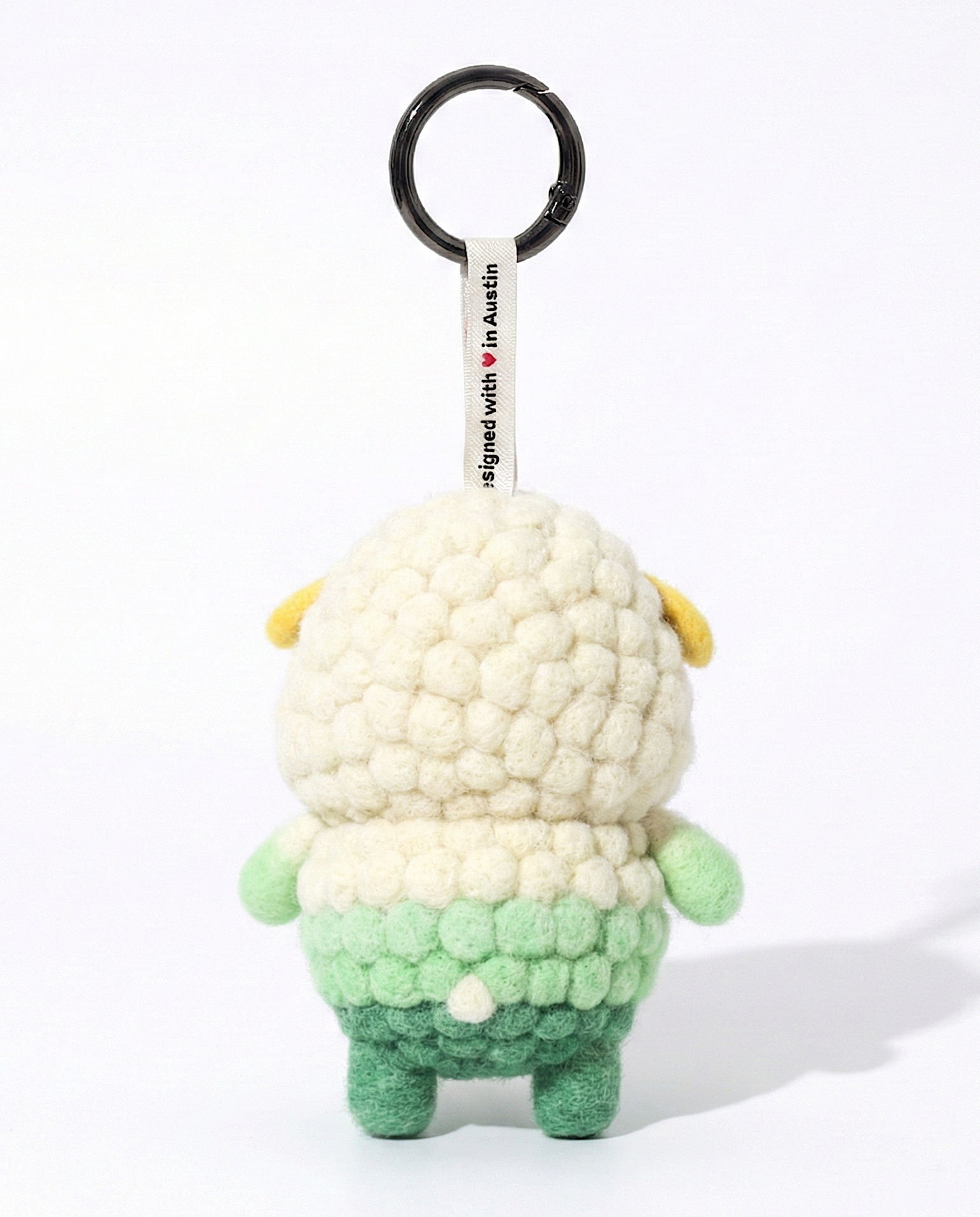 Back view of a handmade Woobbi sheep keychain, made from off-white and green bubbly wool felt. It features a small tag that says 'Designed with ❤️ in Austin' and a round black metal keyring. The sheep has small yellow ears and a tiny white tail. It is set against a plain white background.