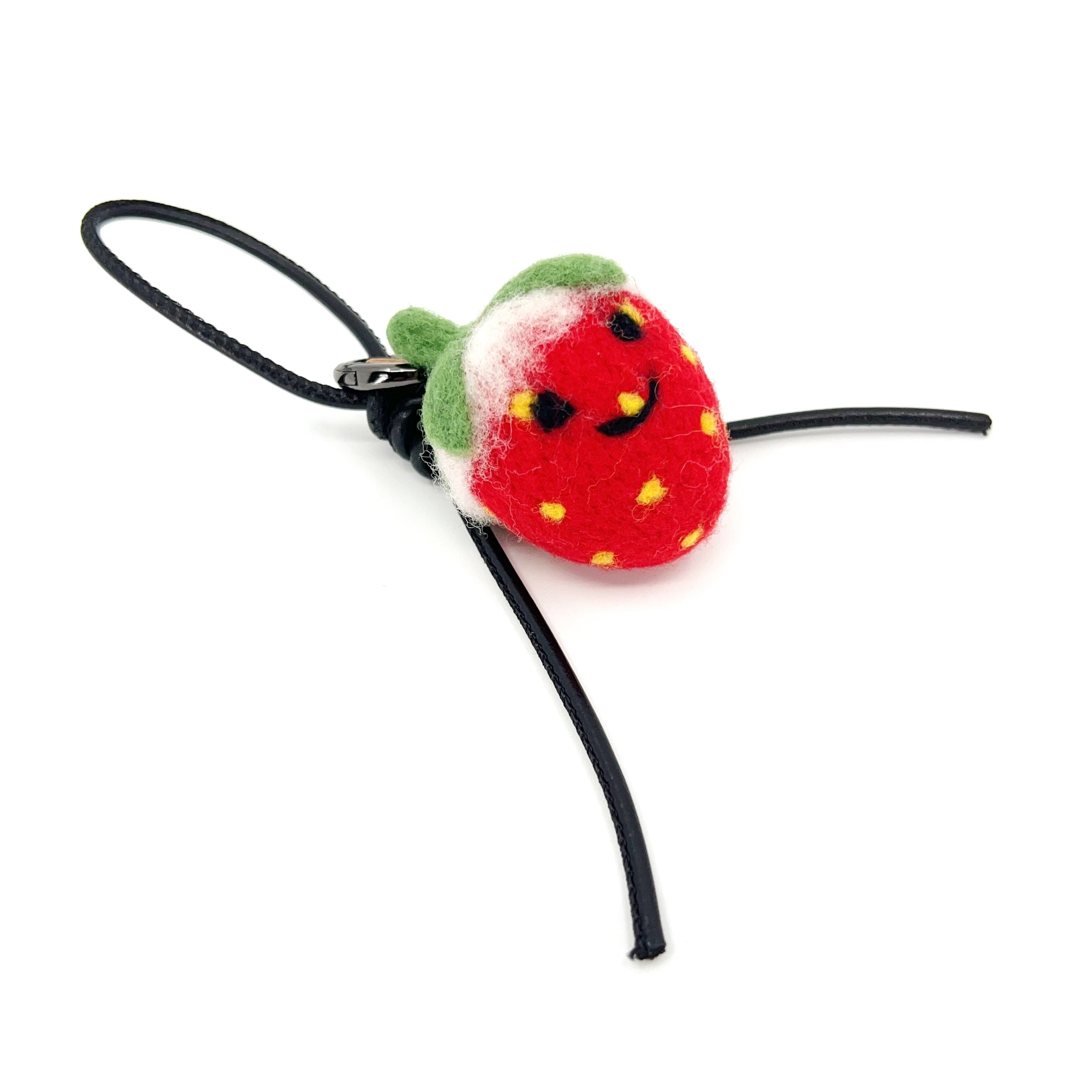Studio shot of needle-felted wool strawberry bag charm featuring a black leather cord and cute smiley face, isolated on a white background.