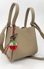 Lifestyle shot of handcrafted needle-felted strawberry bag charm with black leather strap clipped onto a beige leather handbag, showing the cute fruit accessory in use.