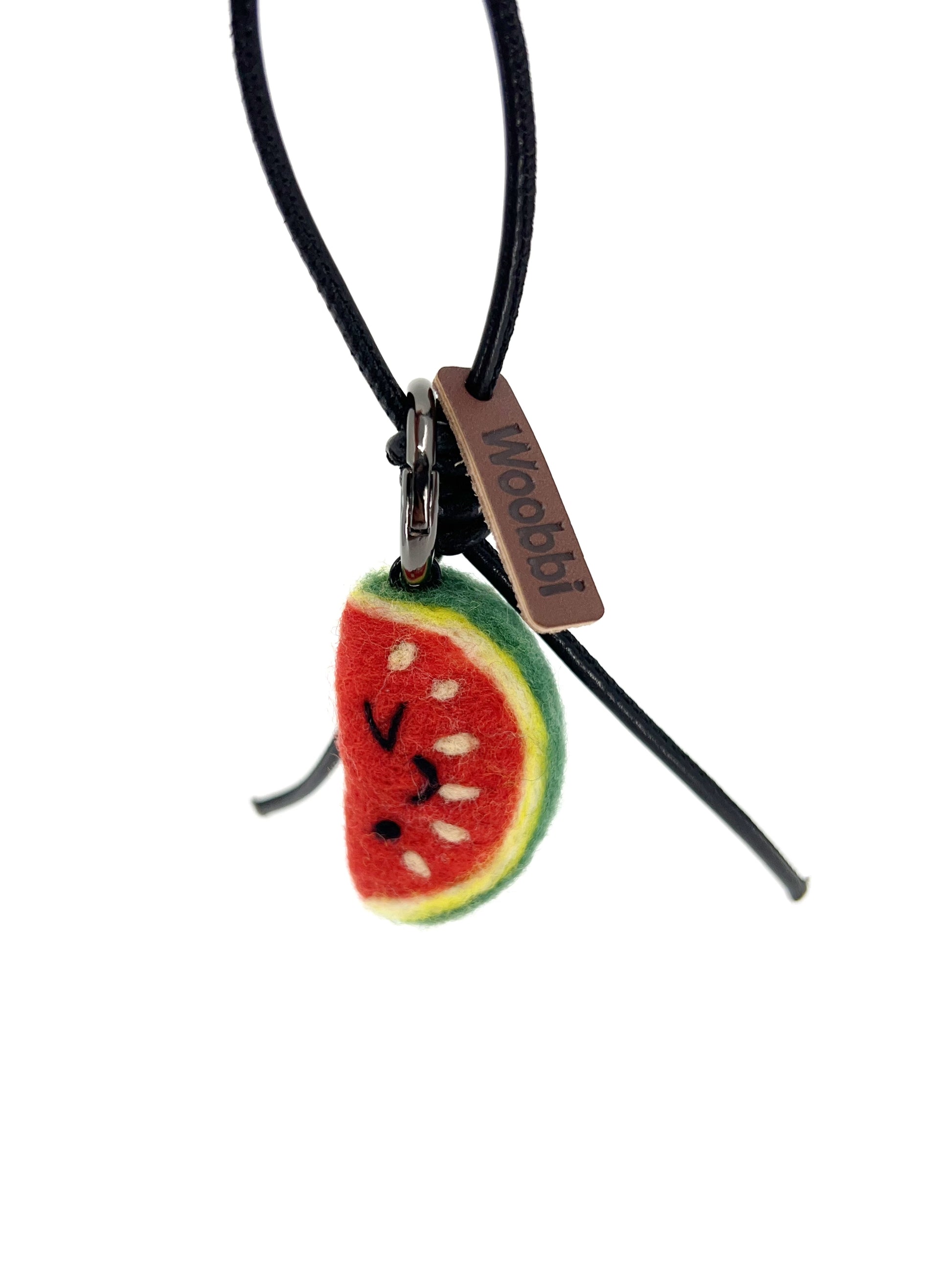 Studio product shot of needle-felted watermelon slice bag charm featuring a black leather cord and brown Woobbi brand tag, isolated on white background.