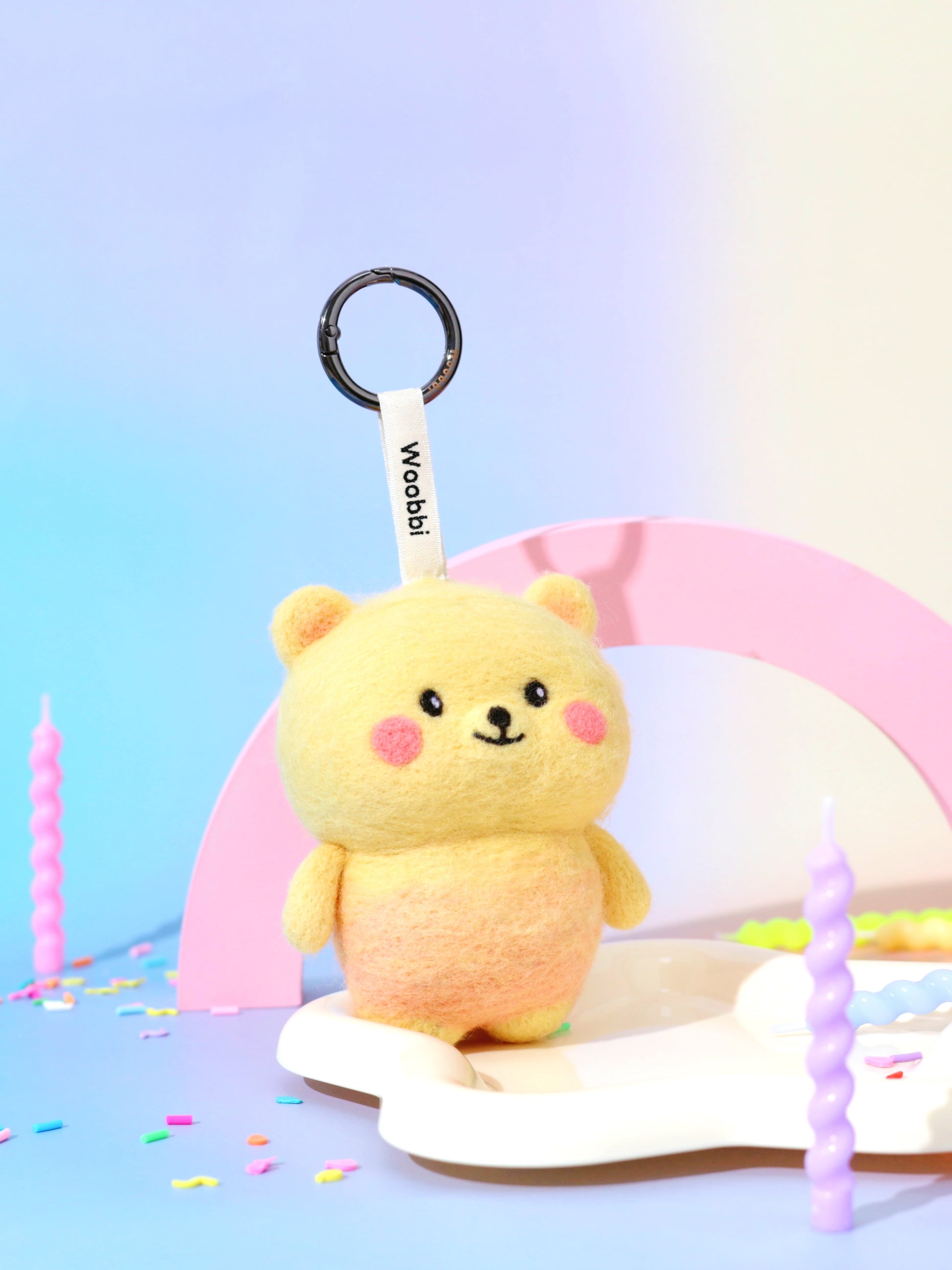 Handcrafted wool bear keychain styled on a pastel party background with colorful candles, showcasing its yellow gradient design and black metal hardware.