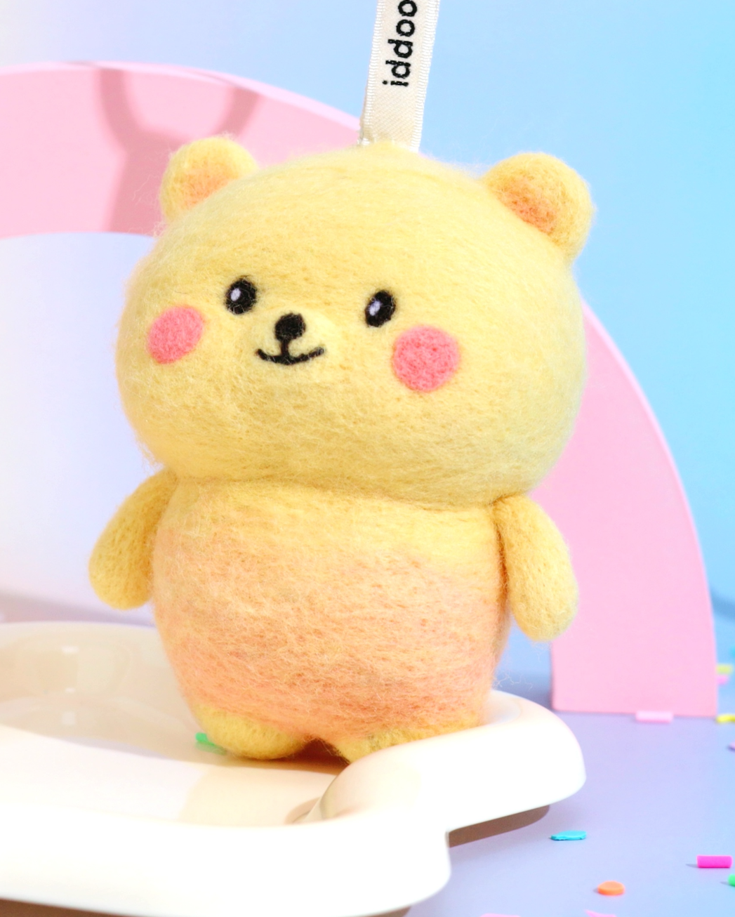Front view of cute yellow needle-felted bear bag charm with a gradient orange belly, pink cheeks, and a white branded Woobbi tag.