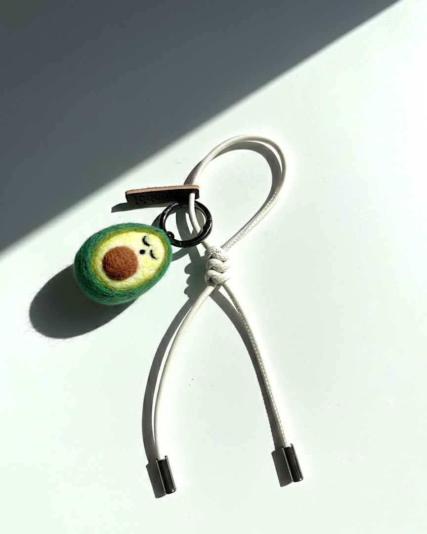Woobbi felted avocado keychain accessory with off-white cream cord on a white background.