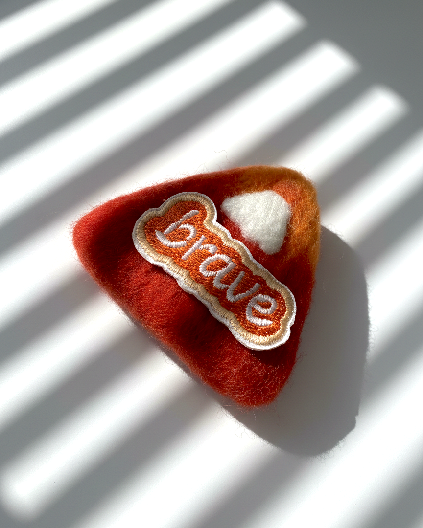 Rust red needle-felted wool magnet with stitched Brave patch, soft rounded shape, handmade fridge decor