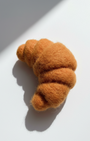 Needle-felted wool croissant magnet shown at an angle with natural light and shadow