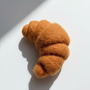 Needle-felted wool croissant magnet shown at an angle with natural light and shadow