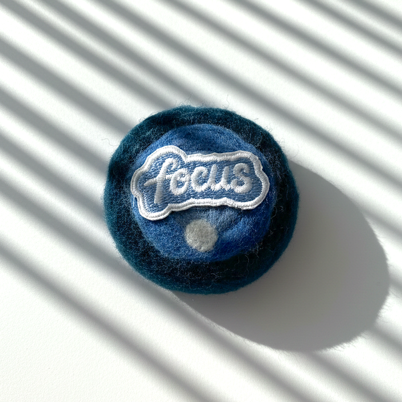Round handmade wool fridge magnet in layered blue tones with embroidered “Focus” patch, needle-felted from merino wool