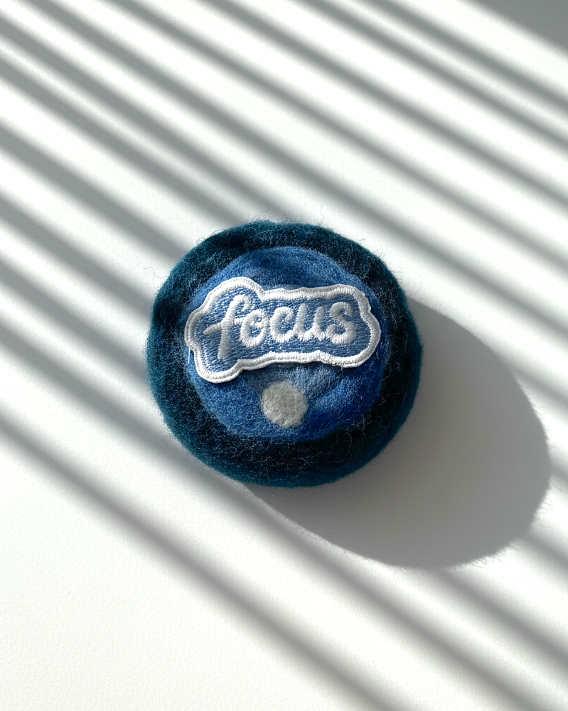 Round handmade wool fridge magnet in layered blue tones with embroidered “Focus” patch, needle-felted from merino wool