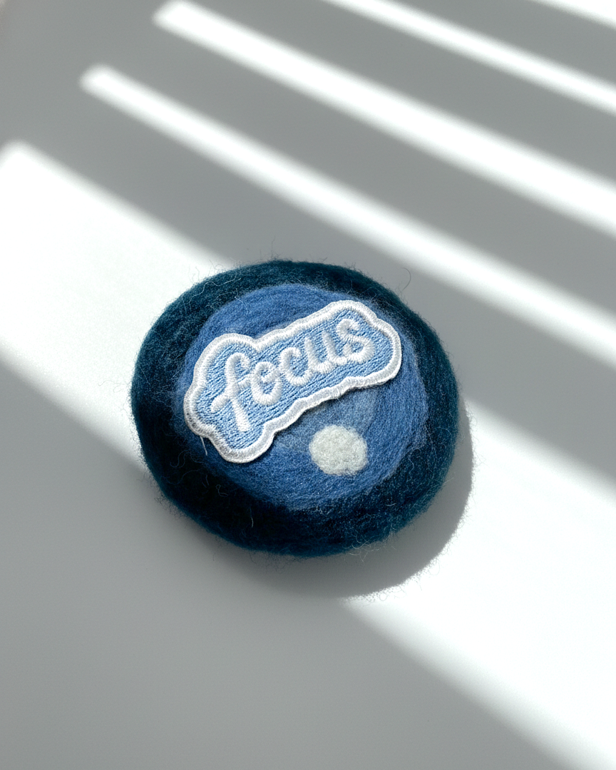 Blue needle-felted wool fridge magnet with stitched Focus patch, round minimalist design made from merino wool