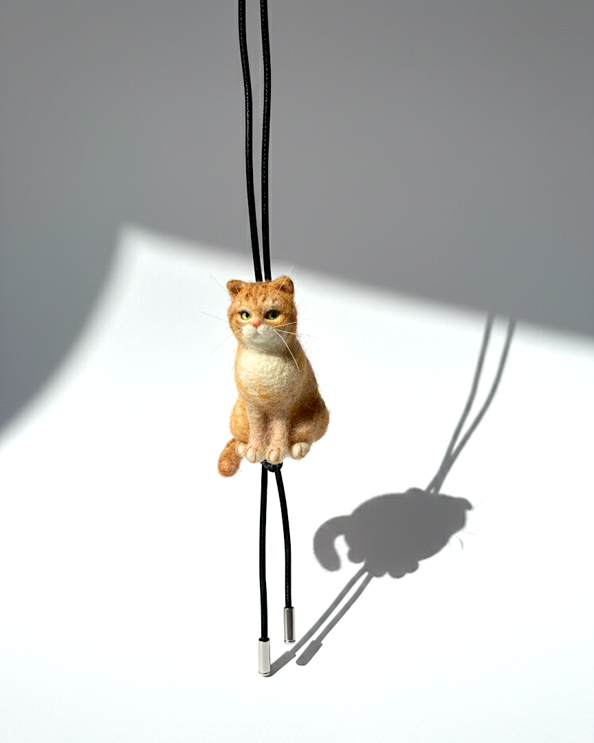 Handmade realistic ginger wool cat sculpture charm in sitting pose, with black sheep skin cord, front view, needle-felted with lifelike details