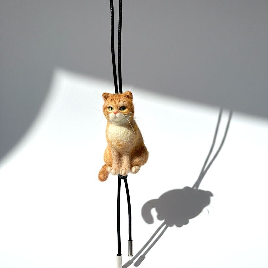 Handmade realistic ginger wool cat sculpture charm in sitting pose, with black sheep skin cord, front view, needle-felted with lifelike details