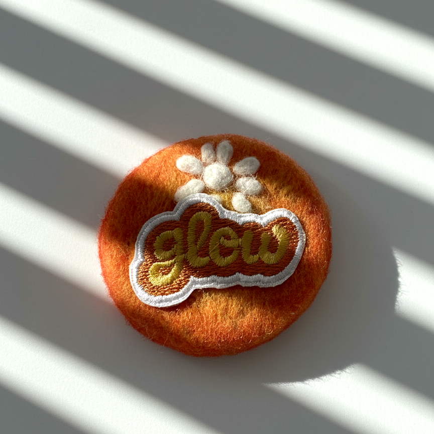 Handmade orange wool fridge magnet with embroidered Glow patch and white flower detail, needle-felted merino wool