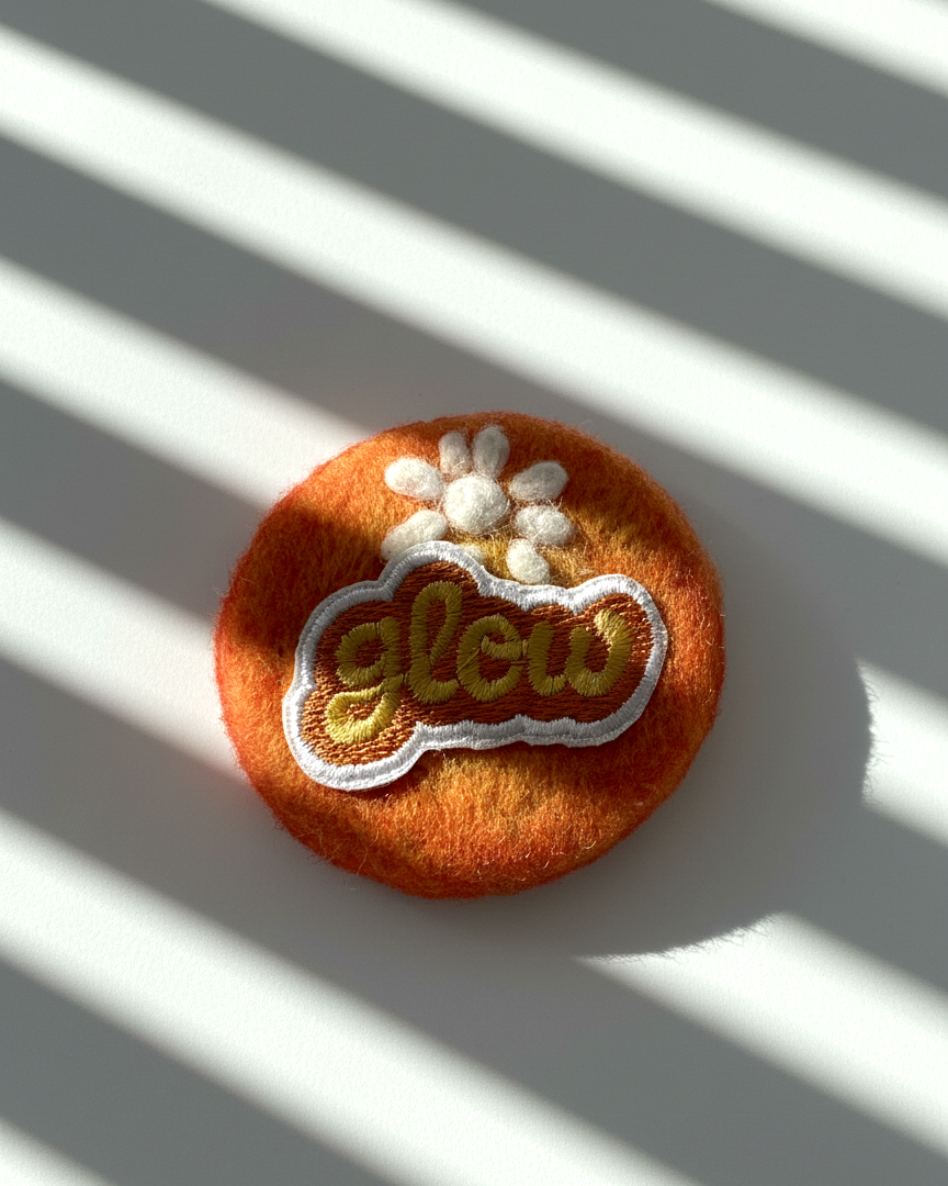 Handmade orange wool fridge magnet with embroidered Glow patch and white flower detail, needle-felted merino wool