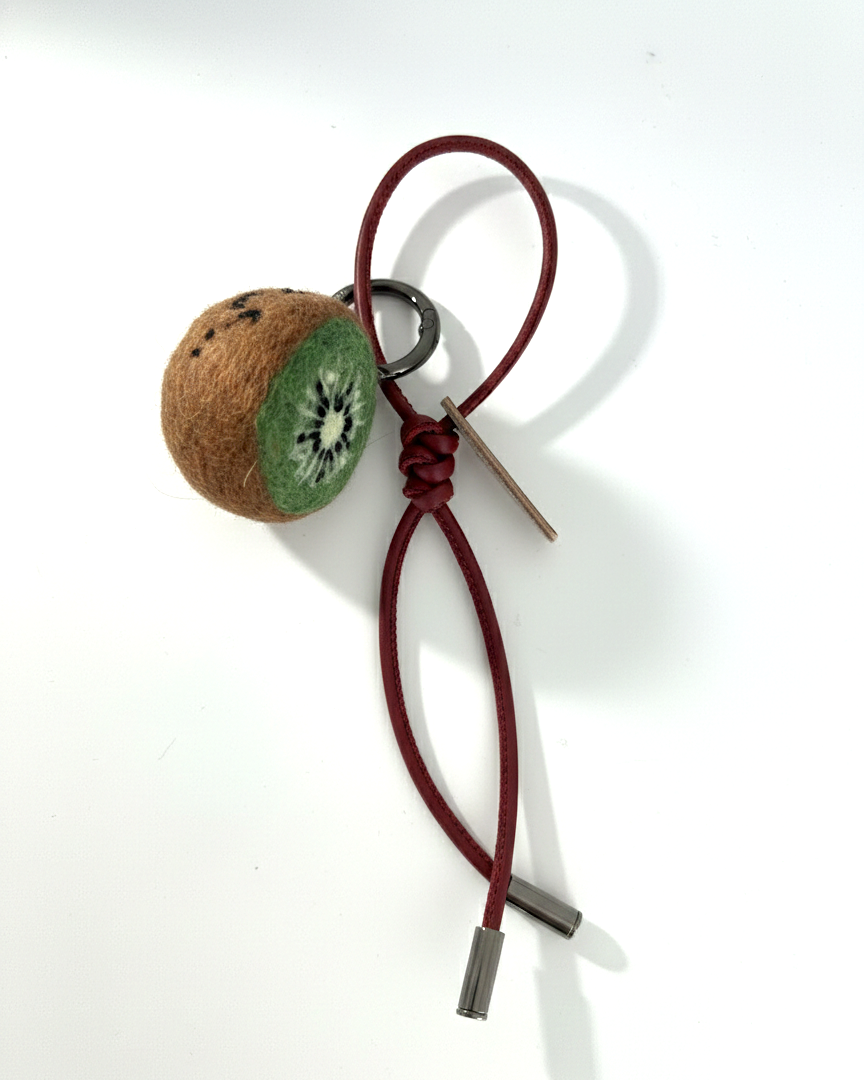 Miniature felted kiwi fruit accessory featuring a dark red leather cord and polished metal tips.