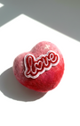 Side view of a 5cm handmade red and pink ombre needle-felted heart magnet featuring a white and red embroidered "Love" patch on a white surface with soft sunlight.