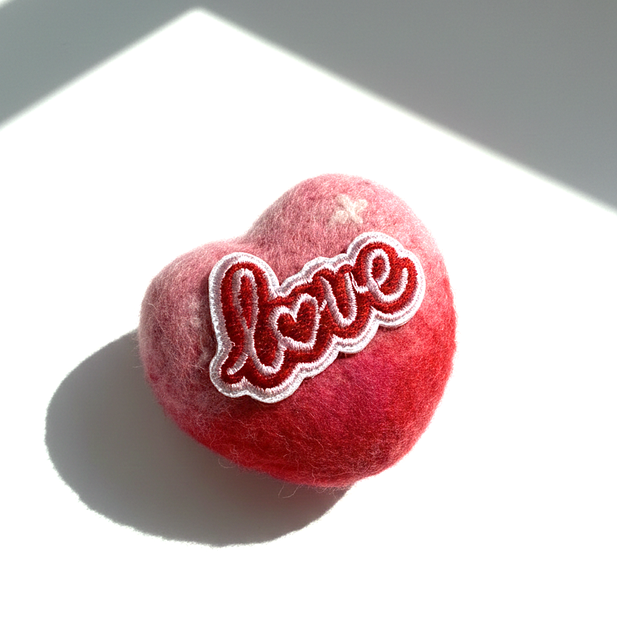 Side view of a 5cm handmade red and pink ombre needle-felted heart magnet featuring a white and red embroidered "Love" patch on a white surface with soft sunlight.