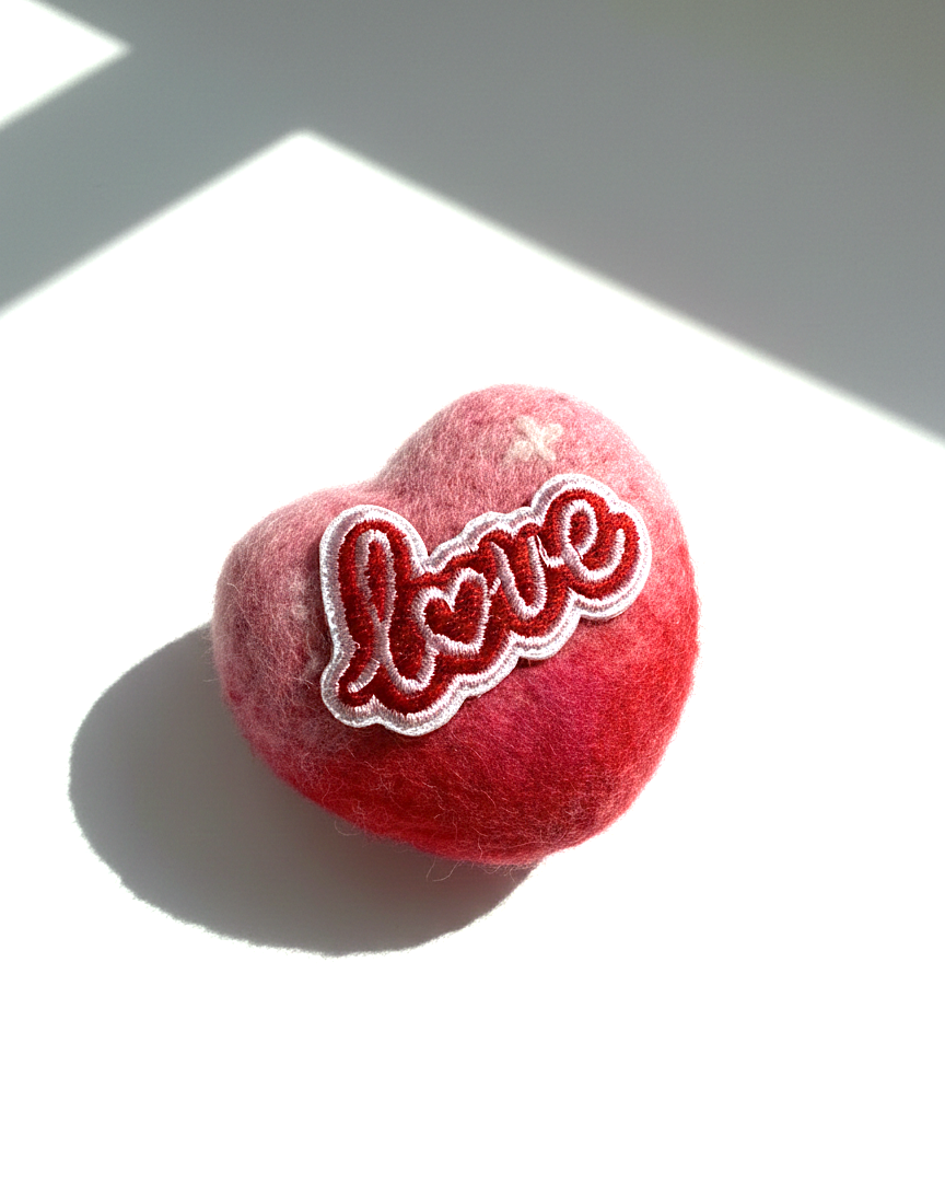Side view of a 5cm handmade red and pink ombre needle-felted heart magnet featuring a white and red embroidered "Love" patch on a white surface with soft sunlight.