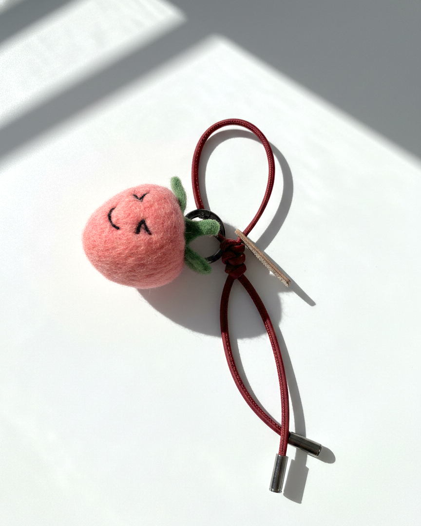 Handmade wool felt strawberry charm with smiling face and red leather loop strap, needle felt fruit bag charm on white background with natural sunlight shadows