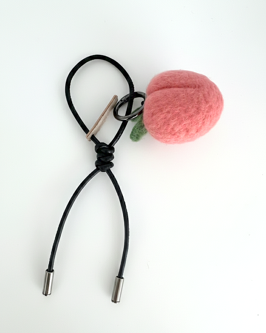 Close-up of handmade wool felt strawberry bag charm with black leather cord and metal tips, sustainable artisan fruit keychain in soft shadow lighting